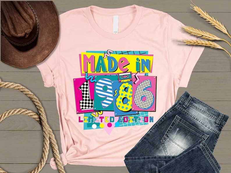 K&ouml;nnte beinhalten: Ein hellrosa T-Shirt mit dem Text "MADE IN 1986 LIMITED EDITION" in einem farbenfrohen Retro-Design. Die Jahreszahl "1986" ist mit verschiedenen Mustern und Farben hervorgehoben. Das Shirt wird mit einer blauen Jeans pr&auml;sentiert.