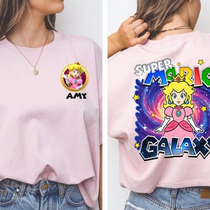 May include: A light pink t-shirt featuring a cartoon image of Princess Peach from Super Mario. The front of the shirt has a circular graphic with the princess and the name "AMY" below. The back of the shirt has the words "SUPER MARIO GALAXY" with a galaxy background.