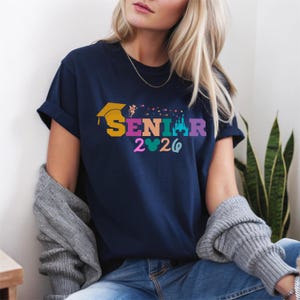 May include: Navy blue t-shirt with the word "SENIOR" in colorful letters, a graduation cap, and the year "2026". The shirt also features a castle and Mickey Mouse ears design. The shirt is a casual style.