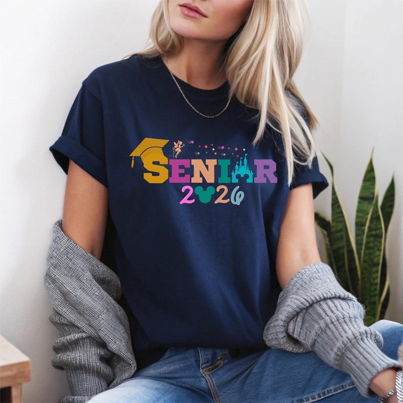 May include: Navy blue t-shirt with the word "SENIOR" in colorful letters, a graduation cap, and the year "2026". The shirt also features a castle and Mickey Mouse ears design. The shirt is a casual style.