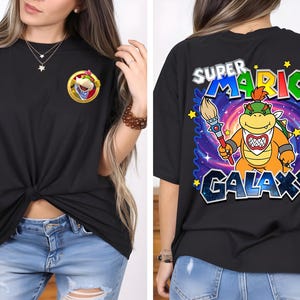 May include: Black t-shirt featuring a Bowser graphic on the front and back. The front graphic is a Bowser character in a gold frame. The back graphic says "Super Mario Galaxy" with a Bowser character holding a paintbrush.