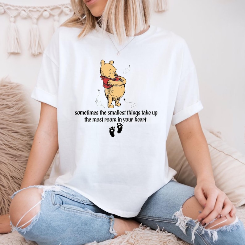 May include: White t-shirt with a cartoon image of Winnie the Pooh hugging himself. The text "sometimes the smallest things take up the most room in your heart" is printed below the image. There are two small footprints below the text.