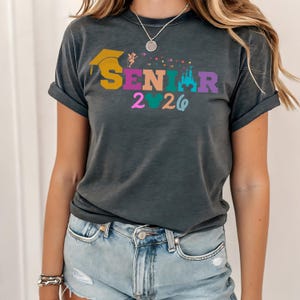 May include: Dark gray t-shirt with rolled sleeves featuring the word "SENIOR" in colorful letters, a graduation cap, a fairy, a castle, and the year "2026". The shirt is worn by a person in light blue denim shorts.