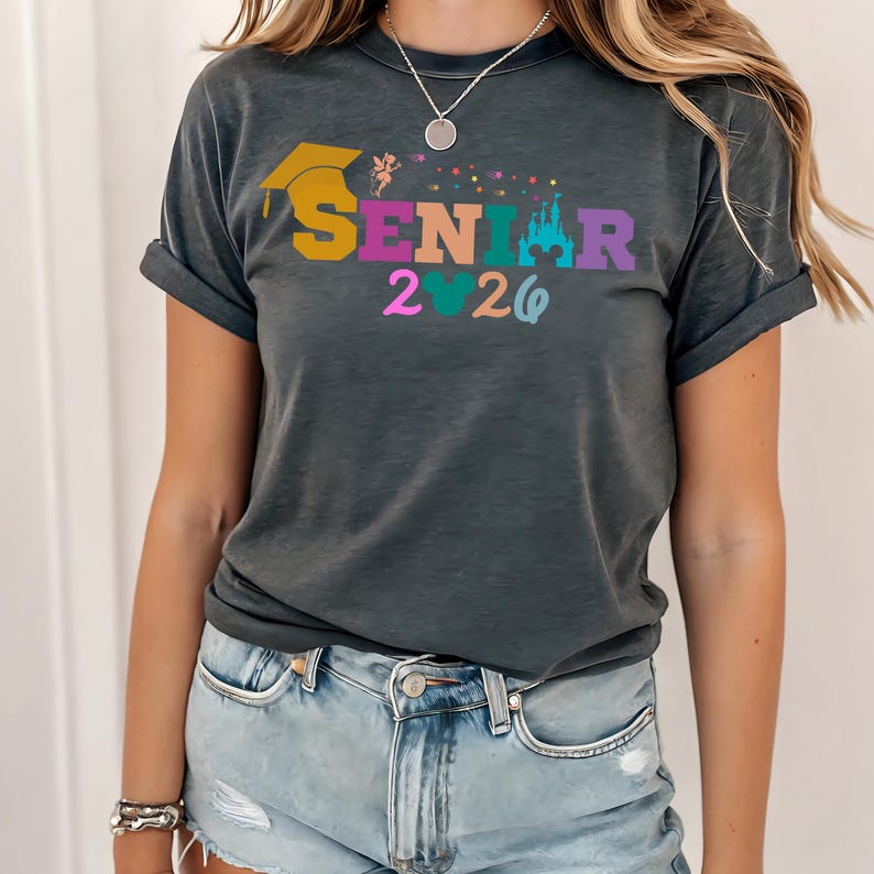 May include: Dark gray t-shirt with rolled sleeves featuring the word "SENIOR" in colorful letters, a graduation cap, a fairy, a castle, and the year "2026". The shirt is worn by a person in light blue denim shorts.