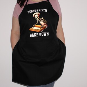May include: Black apron with a skeleton chef rolling out dough. The text on the apron reads "Having a mental bake down."