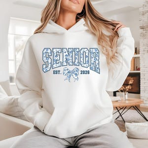 May include: White hooded sweatshirt with the word "SENIOR" in blue floral print. The sweatshirt also features "EST. 2026" and a bow graphic. The hoodie is a comfortable, casual style.