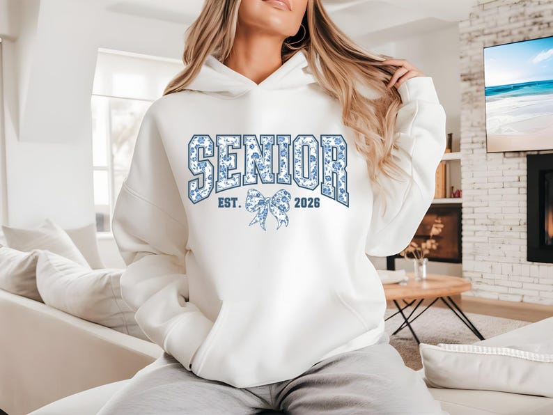 May include: White hooded sweatshirt with the word "SENIOR" in blue floral print. The sweatshirt also features "EST. 2026" and a bow graphic. The hoodie is a comfortable, casual style.