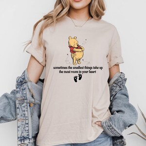 May include: A beige t-shirt with a cartoon image of Winnie the Pooh and the text "sometimes the smallest things take up the most room in your heart".