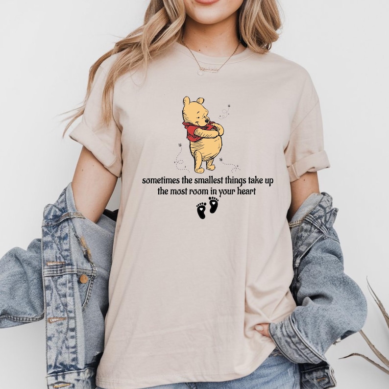 May include: A beige t-shirt with a cartoon image of Winnie the Pooh and the text "sometimes the smallest things take up the most room in your heart".