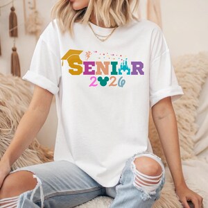 May include: White t-shirt with the word "SENIOR" in colorful letters, a graduation cap, and the year "2026". The design includes a castle and a Mickey Mouse head. The shirt is worn by a person.