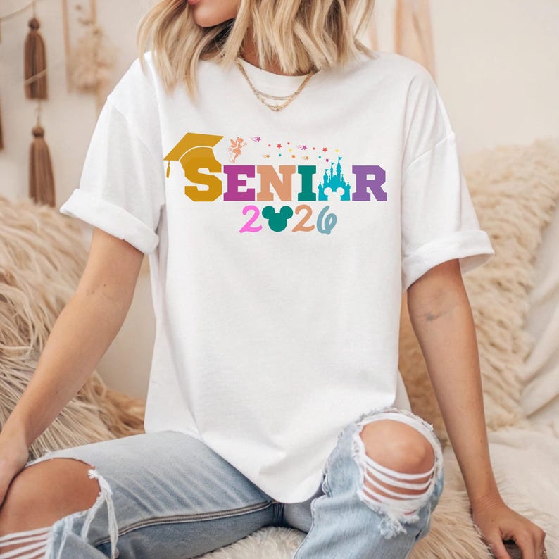 May include: White t-shirt with the word "SENIOR" in colorful letters, a graduation cap, and the year "2026". The design includes a castle and a Mickey Mouse head. The shirt is worn by a person.