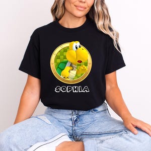 Peut inclure: T-shirt noir avec un personnage de dessin anim&eacute; Koopa Troopa dans un cercle dor&eacute; avec un fond &agrave; carreaux vert et jaune. Le nom "SOPHIA" est imprim&eacute; en blanc en dessous. Le t-shirt est fait d'une mati&egrave;re douce.