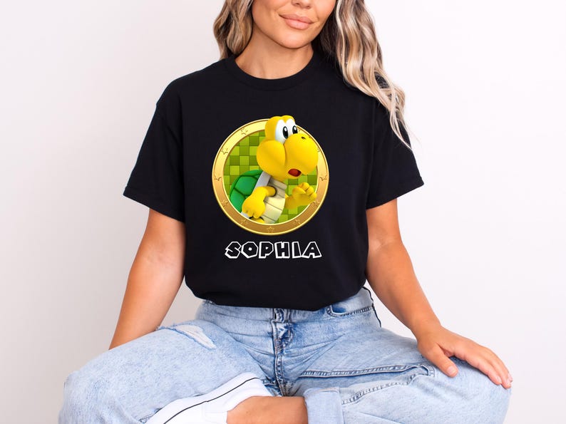 Peut inclure: T-shirt noir avec un personnage de dessin anim&eacute; Koopa Troopa dans un cercle dor&eacute; avec un fond &agrave; carreaux vert et jaune. Le nom "SOPHIA" est imprim&eacute; en blanc en dessous. Le t-shirt est fait d'une mati&egrave;re douce.