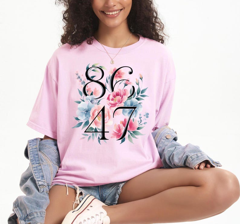 May include: A light pink t-shirt featuring the numbers 86 and 47 in black, surrounded by a floral design with pink and blue flowers and green leaves. The shirt is made of a soft material.