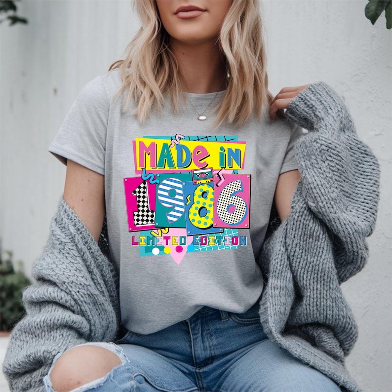 K&ouml;nnte beinhalten: Hellgraues T-Shirt mit einem farbenfrohen Grafikdesign mit der Aufschrift "MADE IN 1986 LIMITED EDITION". Die Grafik zeigt die Zahlen 1, 9, 8 und 6 in verschiedenen Mustern und Farben, im Retro-80er-Jahre-Design.