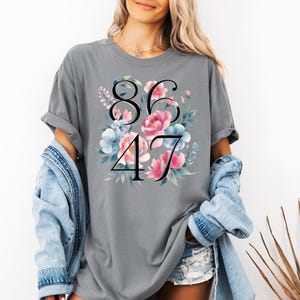 May include: Gray t-shirt with the numbers "8647" in black, surrounded by a floral design with pink and blue flowers. The shirt has short sleeves and a relaxed fit.