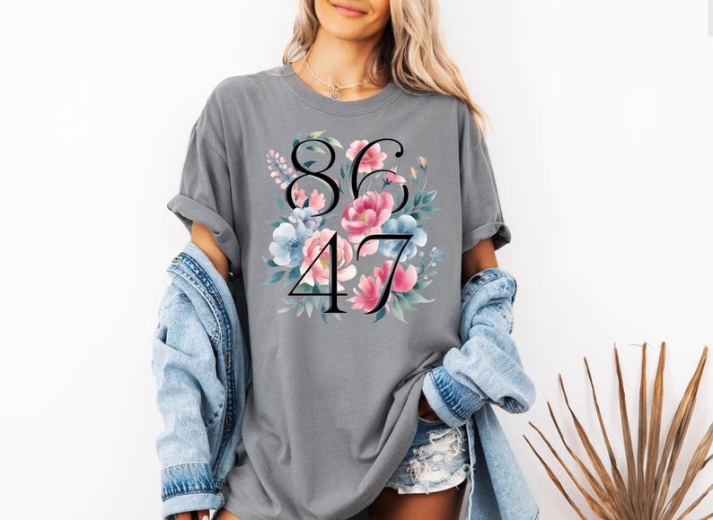 May include: Gray t-shirt with the numbers "8647" in black, surrounded by a floral design with pink and blue flowers. The shirt has short sleeves and a relaxed fit.