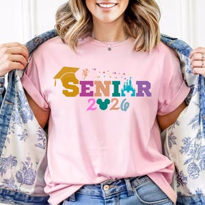 May include: A light pink t-shirt with the word "SENIOR" in colorful letters, a graduation cap, a castle, and the year "2026". The shirt is being worn with a denim jacket.