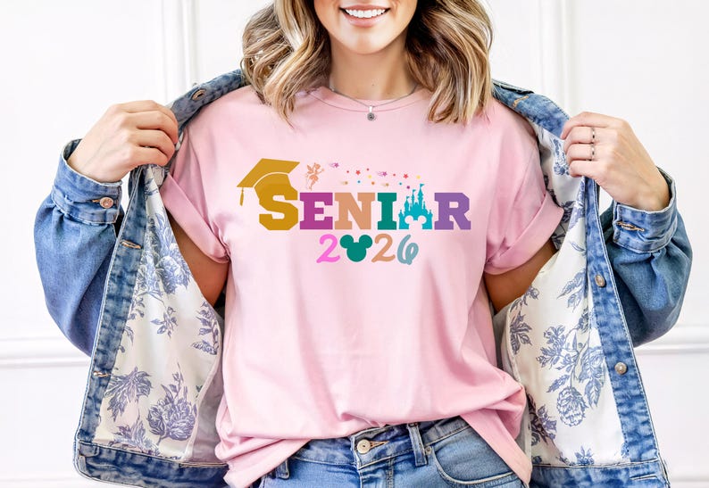 May include: A light pink t-shirt with the word "SENIOR" in colorful letters, a graduation cap, a castle, and the year "2026". The shirt is being worn with a denim jacket.