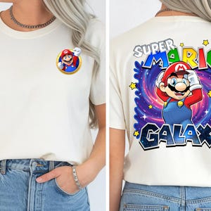 May include: Off-white t-shirt featuring a colorful graphic of Mario from Super Mario Galaxy. The front has a small Mario graphic, while the back displays a larger design with the text "SUPER MARIO GALAXY" and star accents. The shirt is paired with blue jeans.