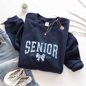 May include: Navy blue sweatshirt with the word "SENIOR" in white letters with a blue floral pattern. The sweatshirt also has the text "EST. 2026" and a bow graphic. A gold necklace and white sneakers are also visible.