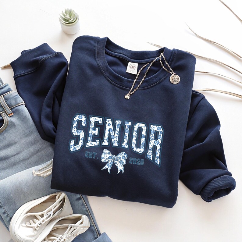 May include: Navy blue sweatshirt with the word "SENIOR" in white letters with a blue floral pattern. The sweatshirt also has the text "EST. 2026" and a bow graphic. A gold necklace and white sneakers are also visible.