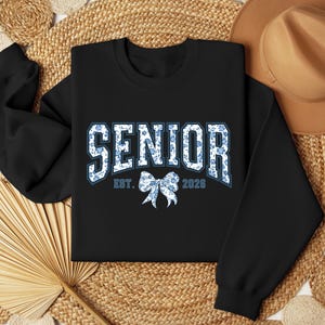 May include: Black sweatshirt with the word "SENIOR" in white and blue floral print lettering. Below the word "SENIOR" is a blue floral bow and the text "EST. 2028". The sweatshirt is displayed on a woven background with a brown hat.