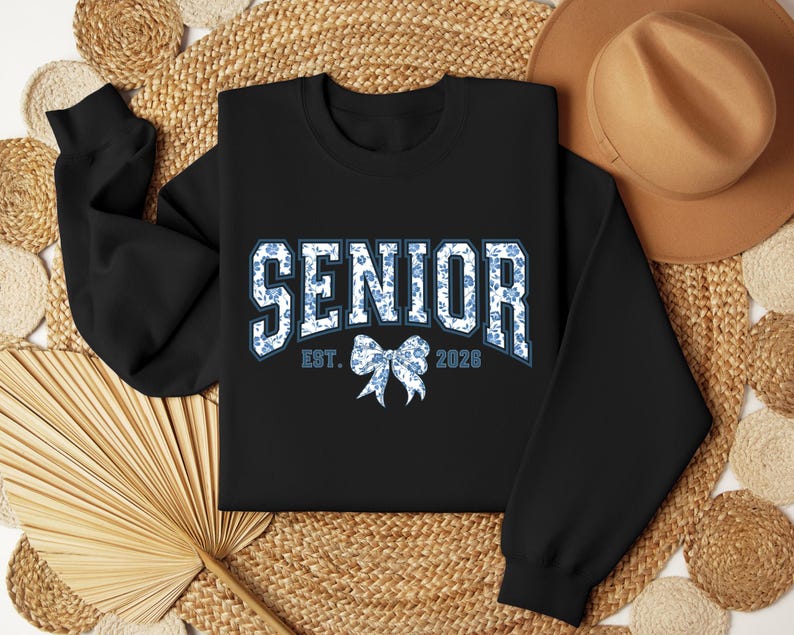 May include: Black sweatshirt with the word "SENIOR" in white and blue floral print lettering. Below the word "SENIOR" is a blue floral bow and the text "EST. 2028". The sweatshirt is displayed on a woven background with a brown hat.