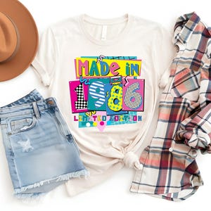 K&ouml;nnte beinhalten: Ein cremefarbenes T-Shirt mit dem Text "MADE IN 1986 LIMITED EDITION" in einem farbenfrohen Retro-Design. Das Shirt ist mit Jeansshorts, einem karierten Hemd und einem braunen Fedora-Hut gestylt.