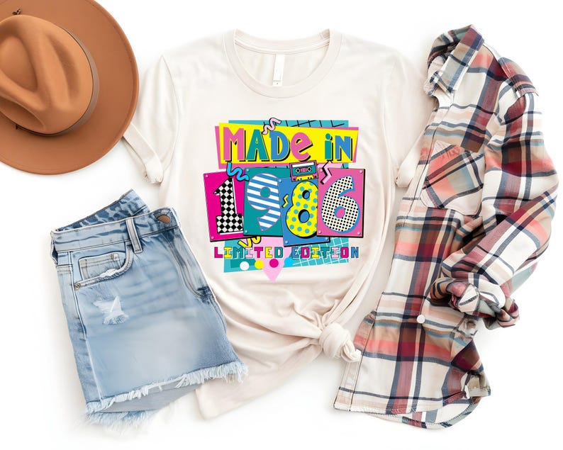 K&ouml;nnte beinhalten: Ein cremefarbenes T-Shirt mit dem Text "MADE IN 1986 LIMITED EDITION" in einem farbenfrohen Retro-Design. Das Shirt ist mit Jeansshorts, einem karierten Hemd und einem braunen Fedora-Hut gestylt.