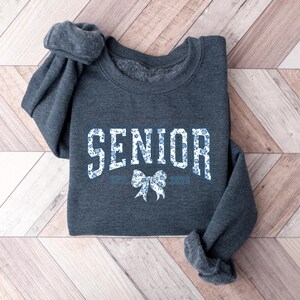 May include: A dark gray sweatshirt with the word "SENIOR" in white block letters filled with a blue floral pattern. Below the word "SENIOR" is a bow graphic and the text "EST. 2025". The sweatshirt is folded on a wooden surface.