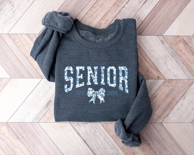 May include: A dark gray sweatshirt with the word "SENIOR" in white block letters filled with a blue floral pattern. Below the word "SENIOR" is a bow graphic and the text "EST. 2025". The sweatshirt is folded on a wooden surface.