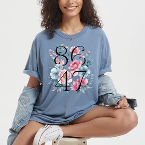 May include: A light blue t-shirt with the numbers 8647 in black, surrounded by a floral design with pink and blue flowers. The shirt has rolled-up sleeves and is worn with a denim jacket.