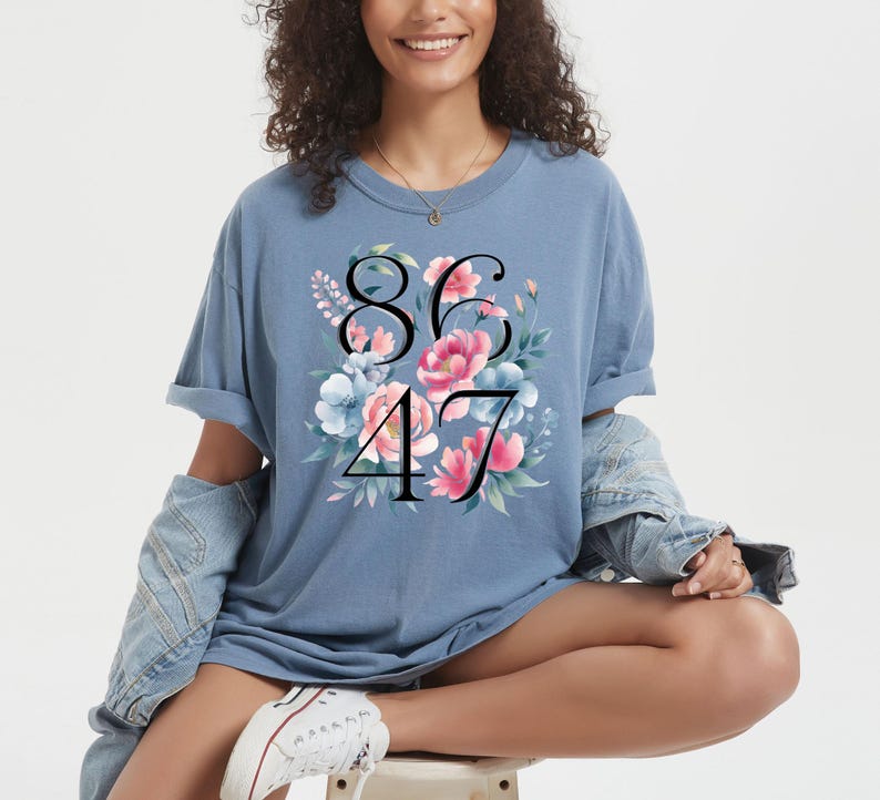 May include: A light blue t-shirt with the numbers 8647 in black, surrounded by a floral design with pink and blue flowers. The shirt has rolled-up sleeves and is worn with a denim jacket.