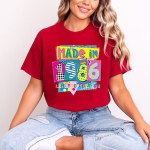 K&ouml;nnte beinhalten: Ein rotes T-Shirt mit einem farbenfrohen Aufdruck mit der Aufschrift "MADE IN 1986 LIMITED EDITION". Die Grafik zeigt die Zahlen 1, 9 und 6 in einem Retro-Stil, mit einer Kassette und einem Schachbrettmuster. Das T-Shirt wird getragen.