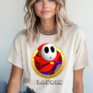 Peut inclure: T-shirt beige avec un personnage de dessin anim&eacute; Shy Guy dans un cercle dor&eacute;. Le personnage porte une tenue rouge et un masque blanc. Le nom "LAUREN" est imprim&eacute; en noir sous le graphisme.