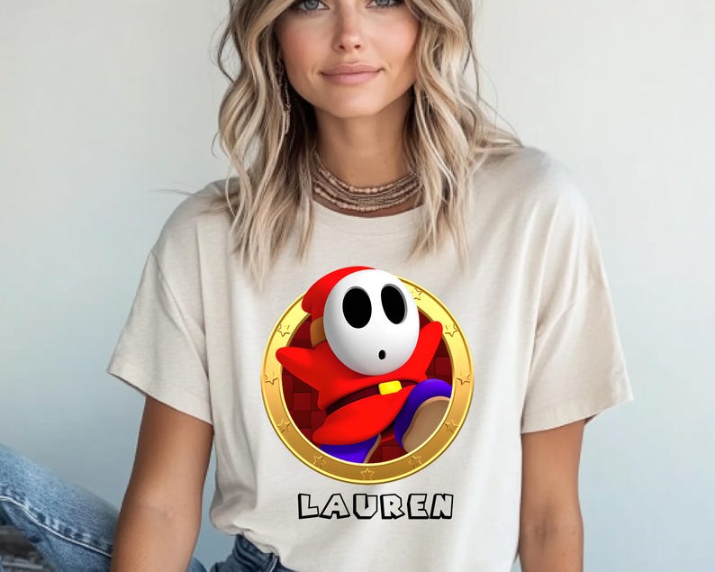 Peut inclure: T-shirt beige avec un personnage de dessin anim&eacute; Shy Guy dans un cercle dor&eacute;. Le personnage porte une tenue rouge et un masque blanc. Le nom "LAUREN" est imprim&eacute; en noir sous le graphisme.