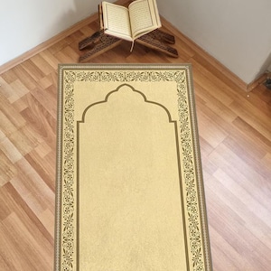 May include: A beige prayer rug with a gold border and a gold arch design. The rug is on a wooden floor.