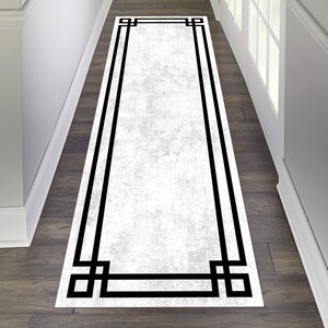 Puede incluir: Una alfombra de pasillo blanca con un borde geométrico negro. La alfombra tiene un aspecto desgastado y texturizado.