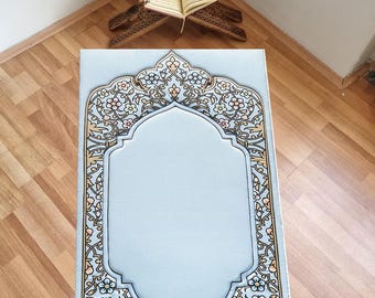 May include: A light blue prayer rug with a floral design in shades of pink, yellow, and brown. The rug has a large, ornate frame around a central rectangle.