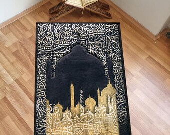 May include: A black prayer rug with a gold mosque design and intricate calligraphy. The rug is laid out on a wooden floor with a wooden prayer stand and an open book on top.