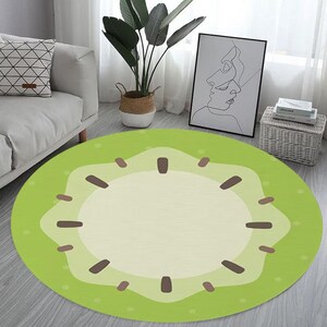 May include: A round green and brown rug with a kiwi fruit design. The rug has a light green background with a darker green kiwi fruit in the center. The kiwi fruit has a brown seed pattern.