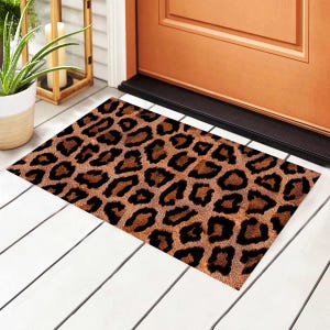 Puede incluir: Una alfombra rectangular con un estampado de leopardo en tonos marrones y negros. La alfombra está colocada frente a una puerta de madera sobre un suelo de madera pintado de blanco.