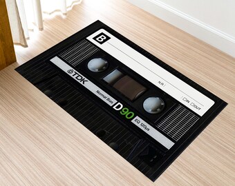 Könnte beinhalten: Schwarze, rechteckige Fußmatte im Design einer TDK D90-Kassette. Die Matte zeigt weißen und silbernen Text, darunter "TDK", "D90" und "Normal Bias". Das Design umfasst ein Rastermuster und einen weißen Etikettenbereich.