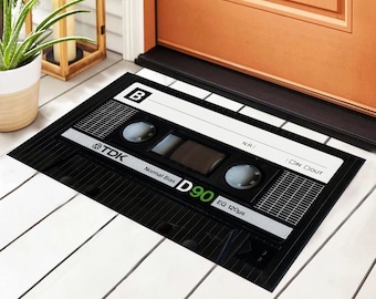 Könnte beinhalten: Schwarze rechteckige Fußmatte mit einem grafischen Design einer Kassette. Die Matte zeigt den Text "TDK", "D90" und weitere Details, mit einem weißen und grauen Farbschema. Die Matte liegt vor einer Holztür.