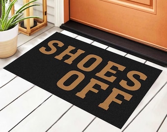 May include: Black rectangular doormat with the words "SHOES OFF" in a bold, tan font. The mat is placed in front of a wooden door on white-painted wooden planks. A potted plant and decorative lantern are visible.