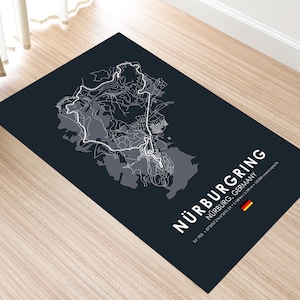May include: A dark blue rectangular rug featuring a detailed white and gray map of the Nürburgring race track in Germany. The text "NÜRBURGRING" is displayed in white, along with the track's location and a German flag.