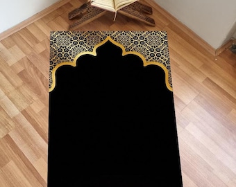 May include: A black prayer rug with a gold intricate design. The rug features a large, gold arch with a smaller, gold arch inside. The rug is surrounded by a gold border with a repeating pattern. The rug is on a wooden floor.