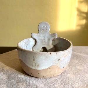 May include: A white ceramic bowl with a brown rim and a white ceramic gingerbread man figurine inside. The bowl has a speckled glaze and a wavy design.