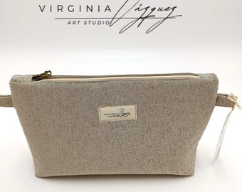 May include: A rectangular, light gray cosmetic bag with a zipper closure and a small label that reads "VIRGINIA JÍZQUEZ ART STUDIO." The bag is made of a textured fabric and has a neutral color palette. A tag is attached to the side.